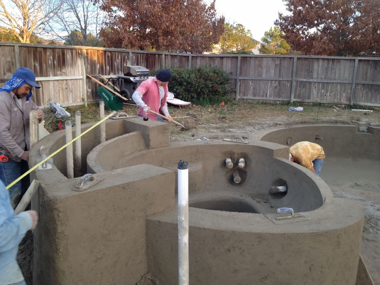 Crew finishing a freeform pool and spa shell in a suburban backyard, plumbing rising from shell floor