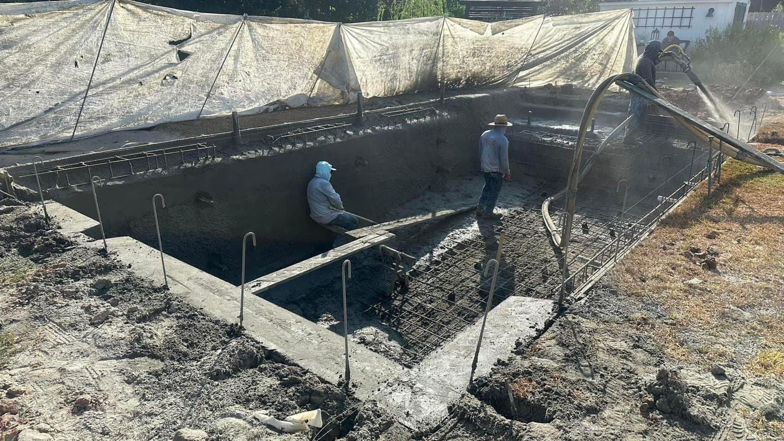 Two workers finishing a partially gunited rectangular pool shell in an open yard