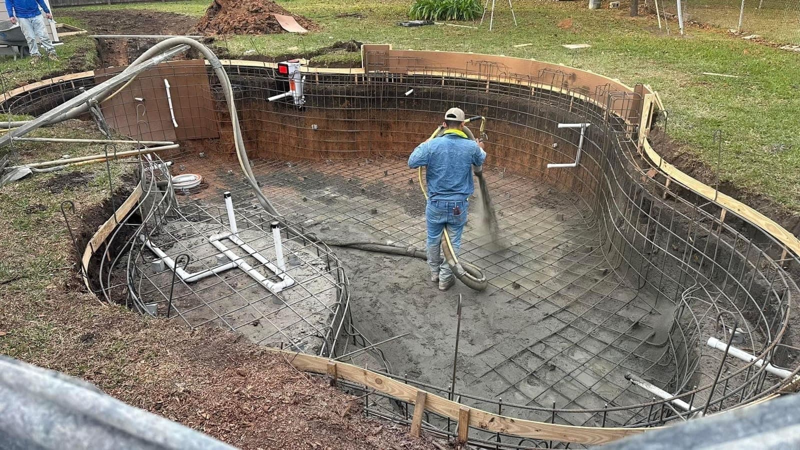Nozzleman spraying gunite from inside an oval pool shell, rebar cage and wooden forms visible