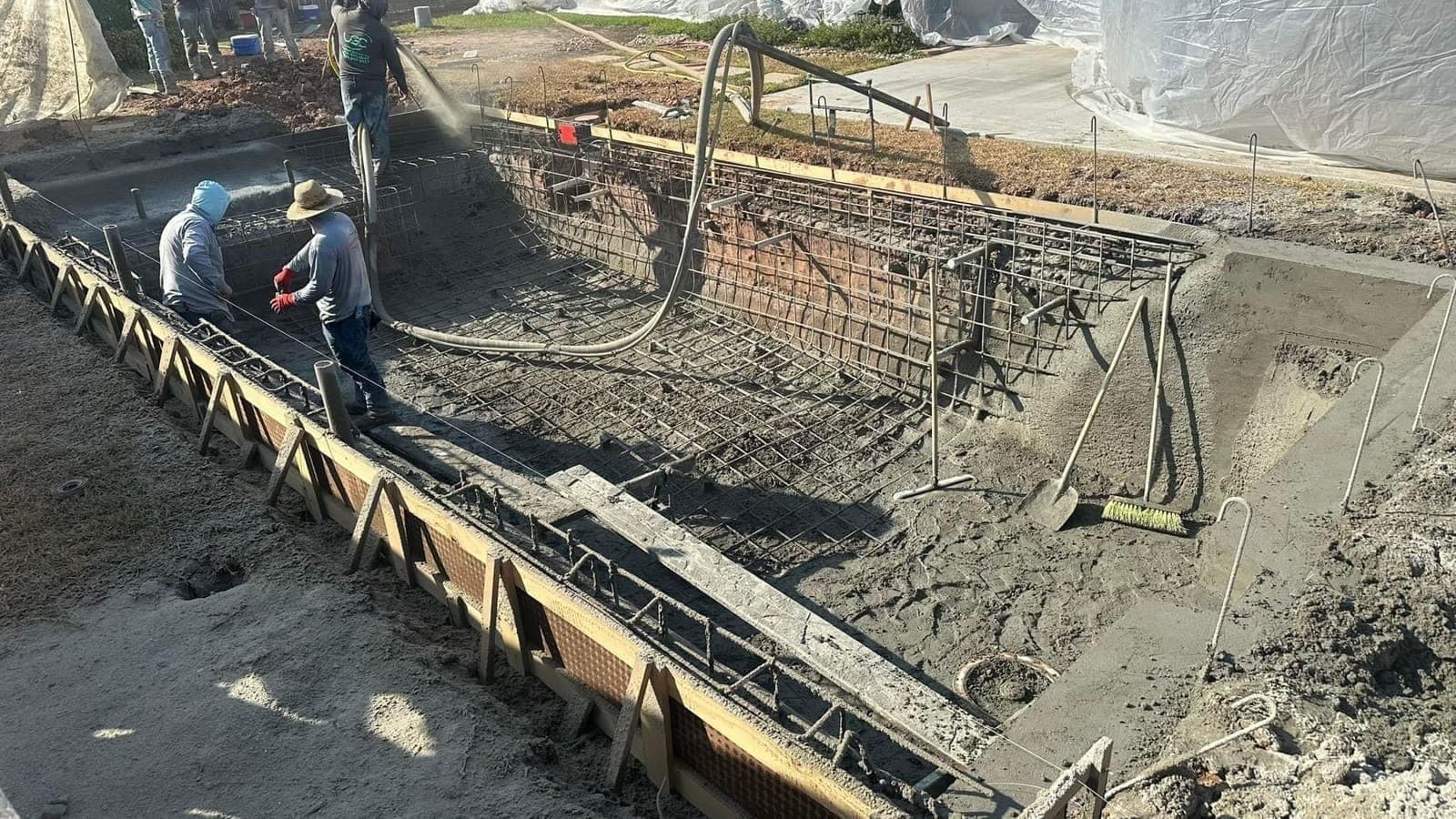 Two workers applying gunite on an L-shaped pool shell with rebar cage and formwork